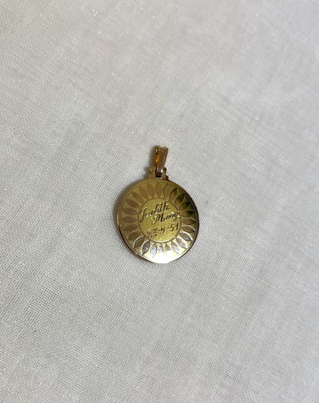 gold disc pendant with name and date engraving, resting on white fabric background 