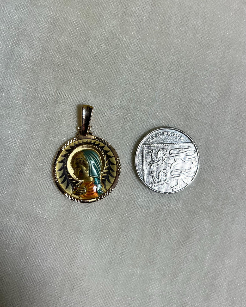 gold pendant with enamel Virgin Mary, next to ten pence coin, on white fabric background