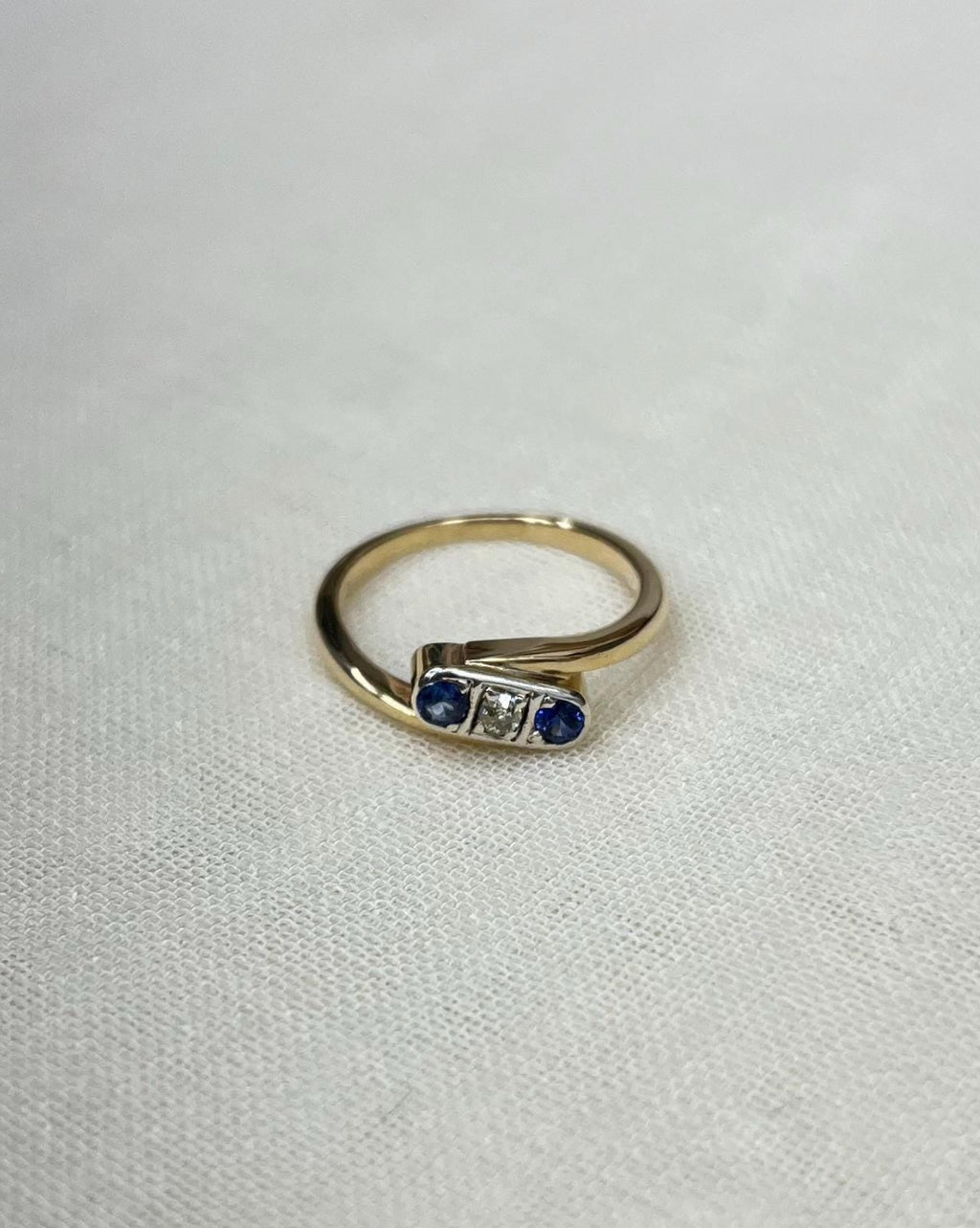 gold trilogy crossover ring with 2 blue sapphire and diamond centre, resting on a white fabric background 