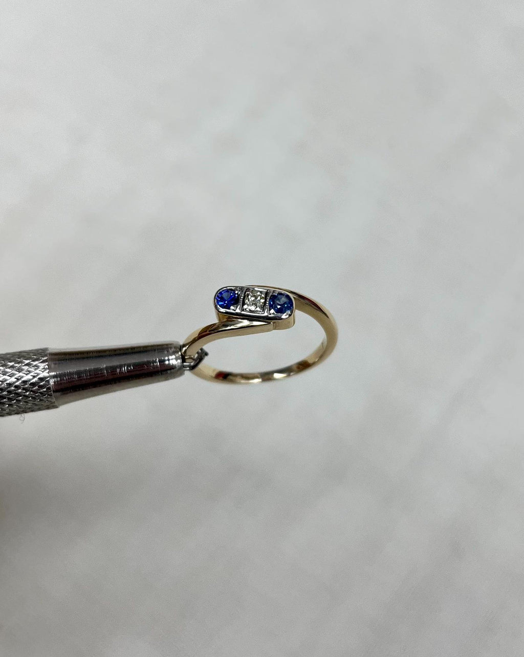 gold trilogy crossover ring with 2 blue sapphires and a diamond, held by metal tool above white background 