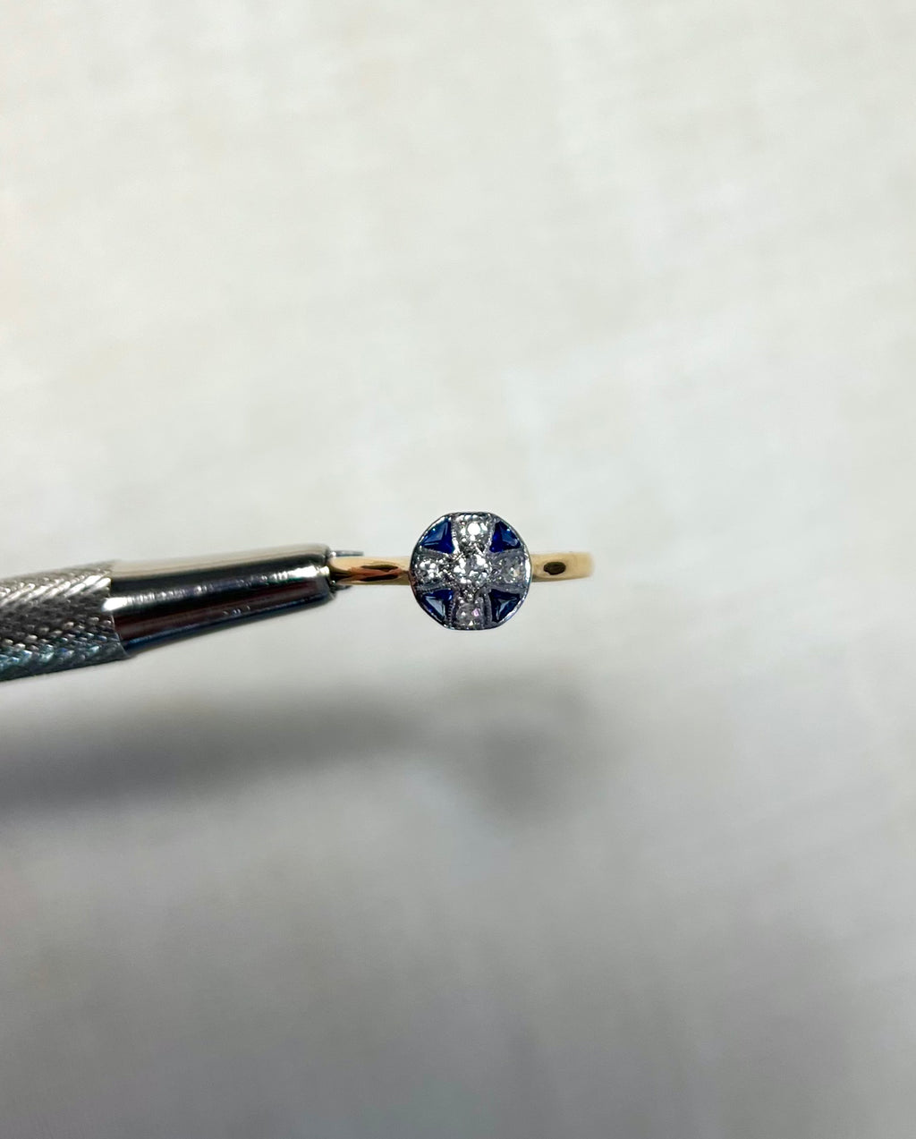 round gold ring with blue sapphire and diamond stones, held by silver tweezers above white background