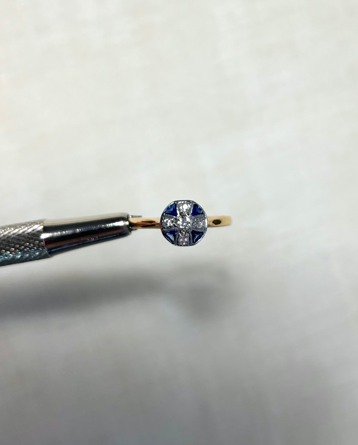 round gold ring with blue sapphire and diamond stones, held by silver tweezers above white background