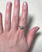 A close-up of a hand wearing a gold ring with a dark blue cross-shaped stone and small clear stones around it, set against a light beige background.