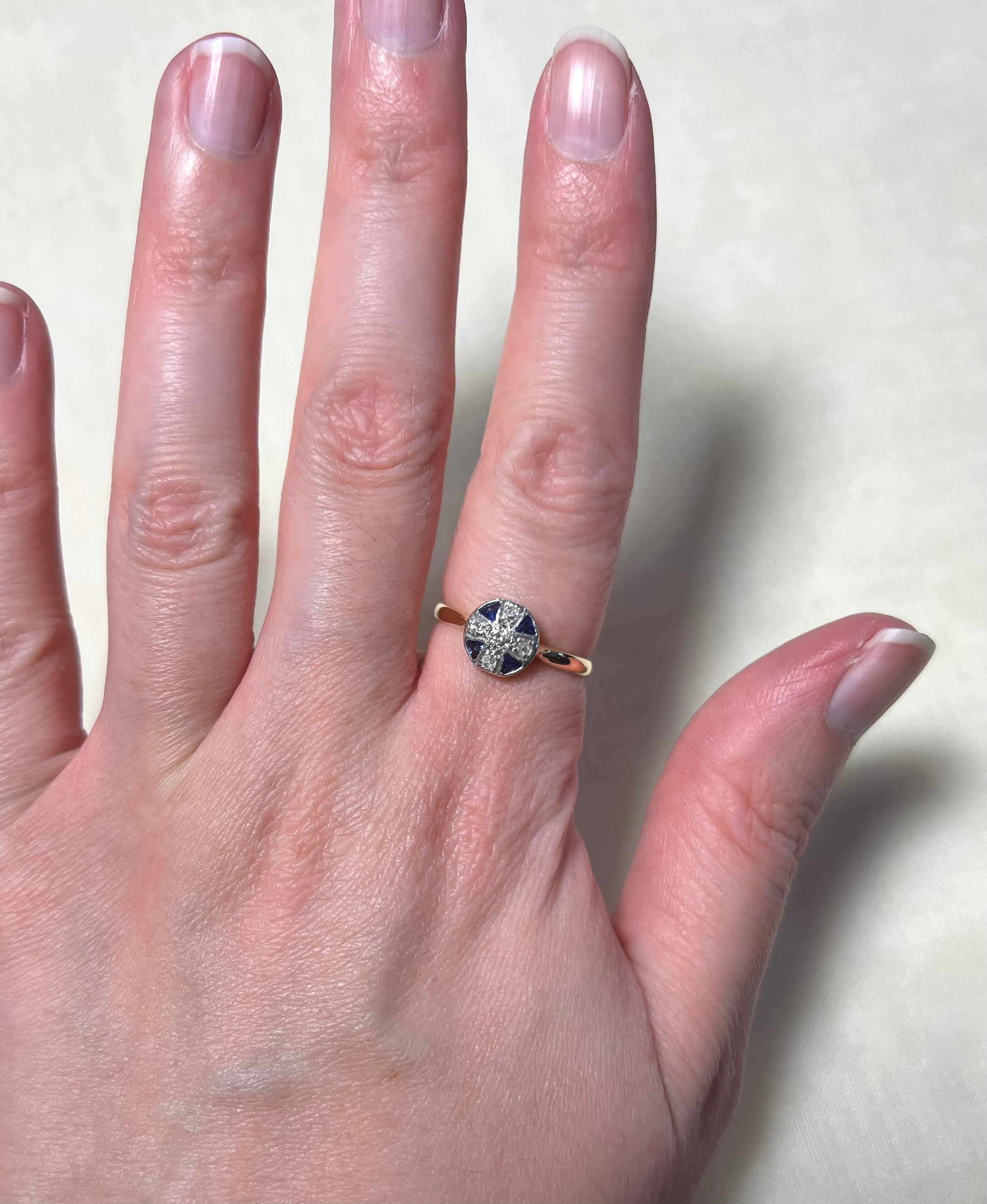A close-up of a hand wearing a gold ring with a dark blue cross-shaped stone and small clear stones around it, set against a light beige background.