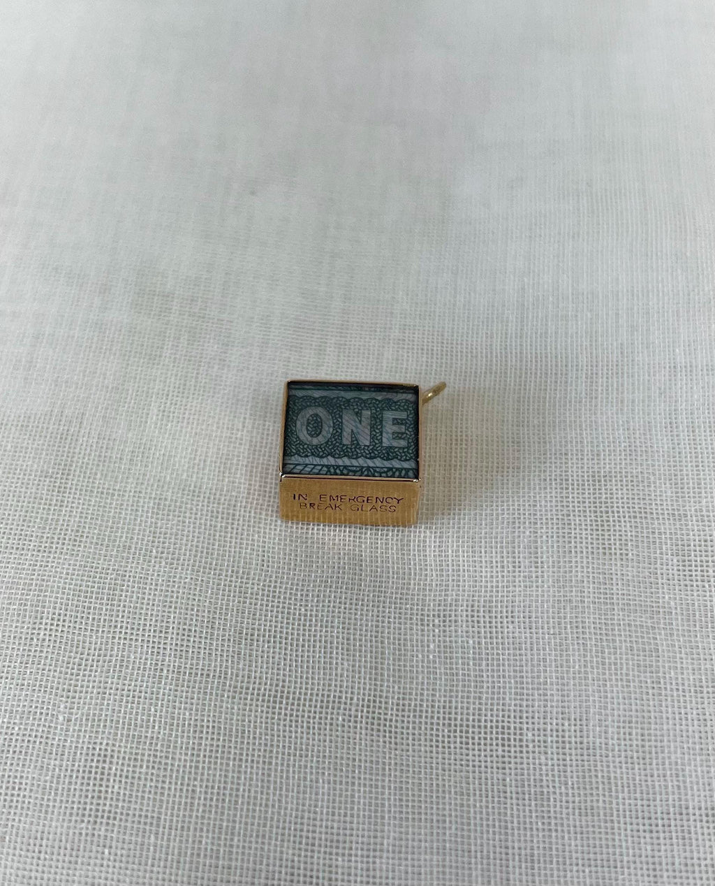 gold charm with one pound note inside and "in emergency break glass" engraving, resting on white fabric background