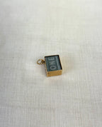 side view of square gold charm containing visible one pound note, resting on white fabric background