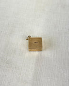 back of square gold charm with visible hallmark and engraving "in emergency break glass" on the bottom, on white fabric background