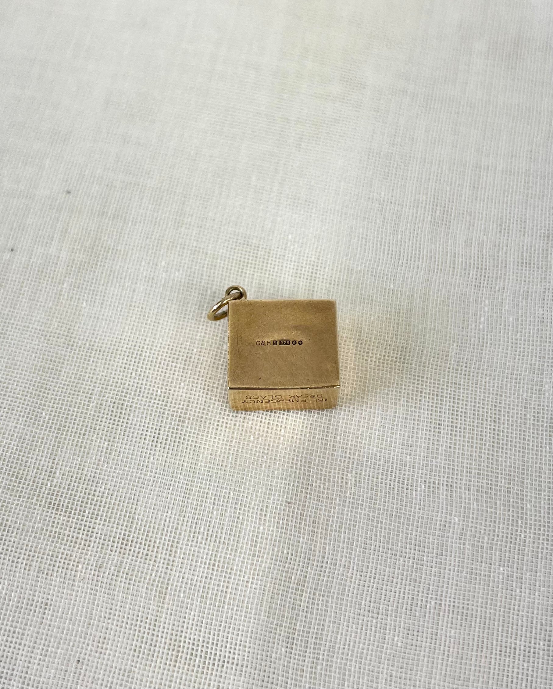 back of square gold charm with visible hallmark and engraving "in emergency break glass" on the bottom, on white fabric background