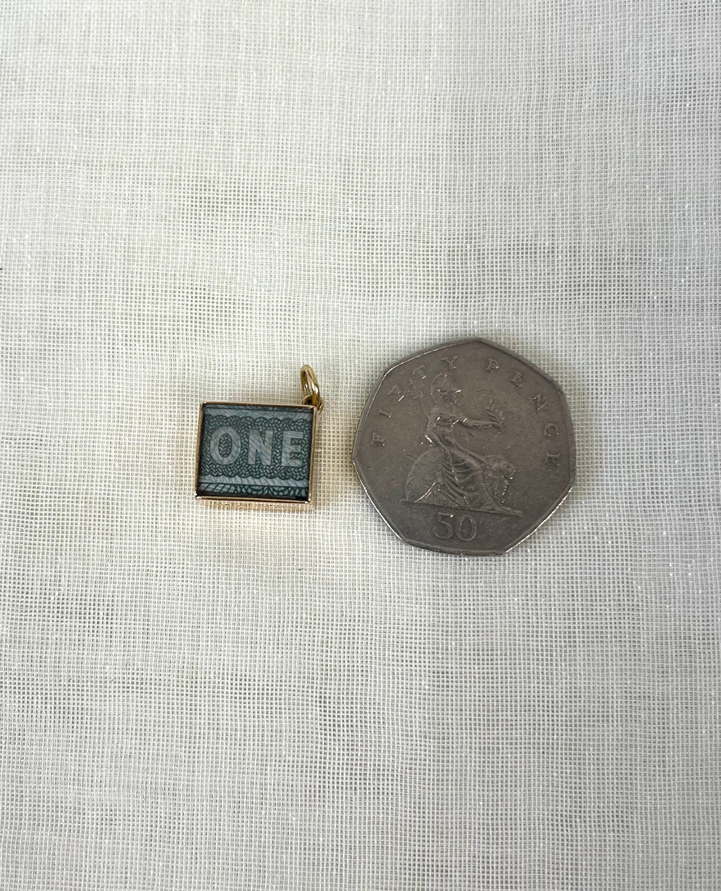 square gold charm with one pound note inside next to fifty pence coin for size reference, sitting on white fabric background.