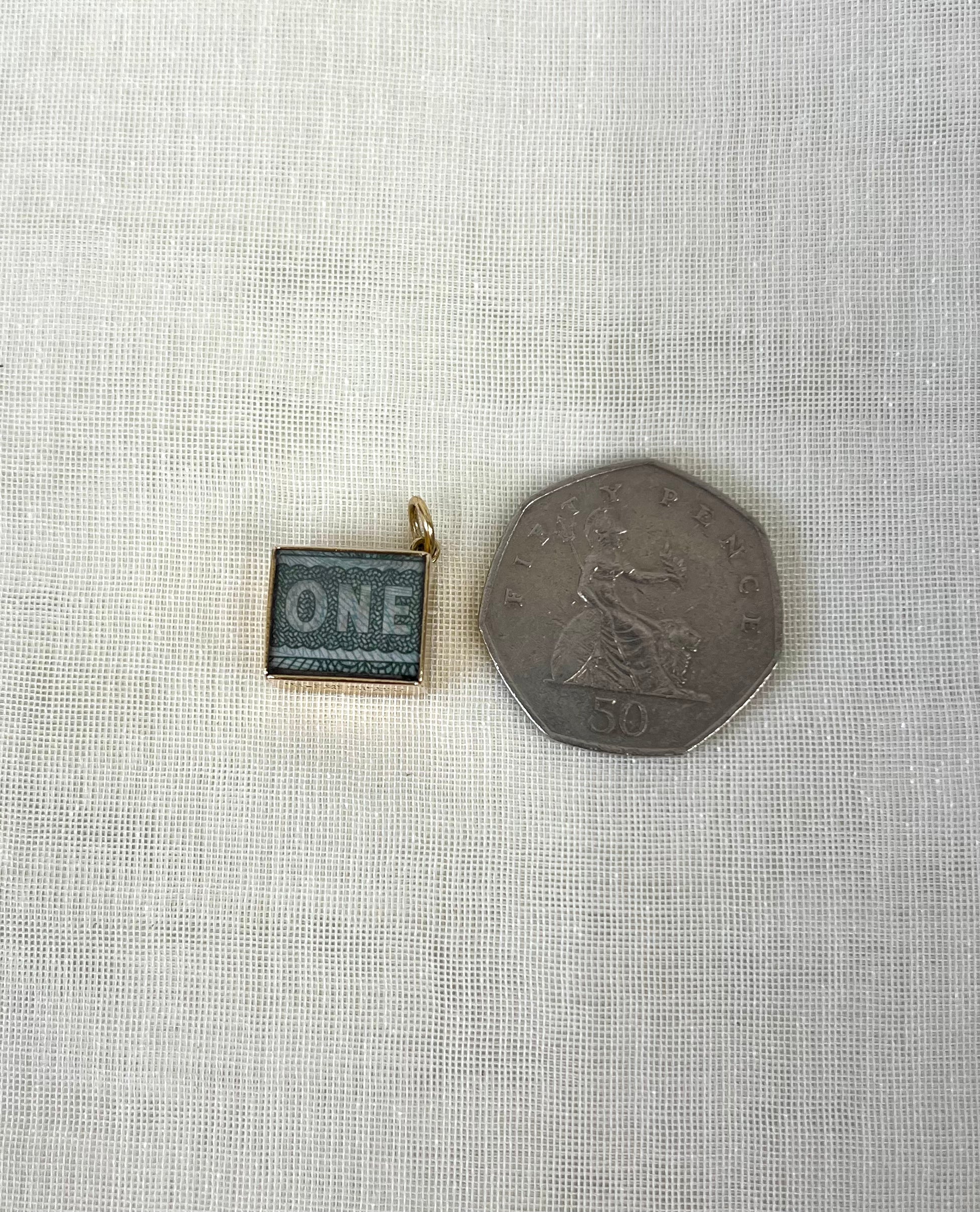 square gold charm with one pound note inside next to fifty pence coin for size reference, sitting on white fabric background.