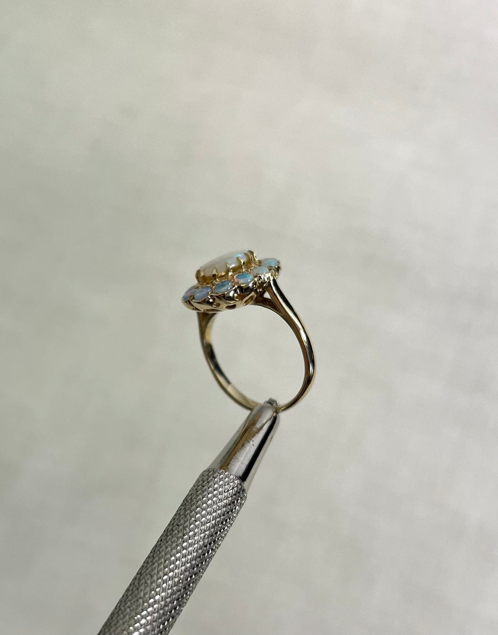 side view of gold ring with multiple opal stones, help by silver metal tool above white background