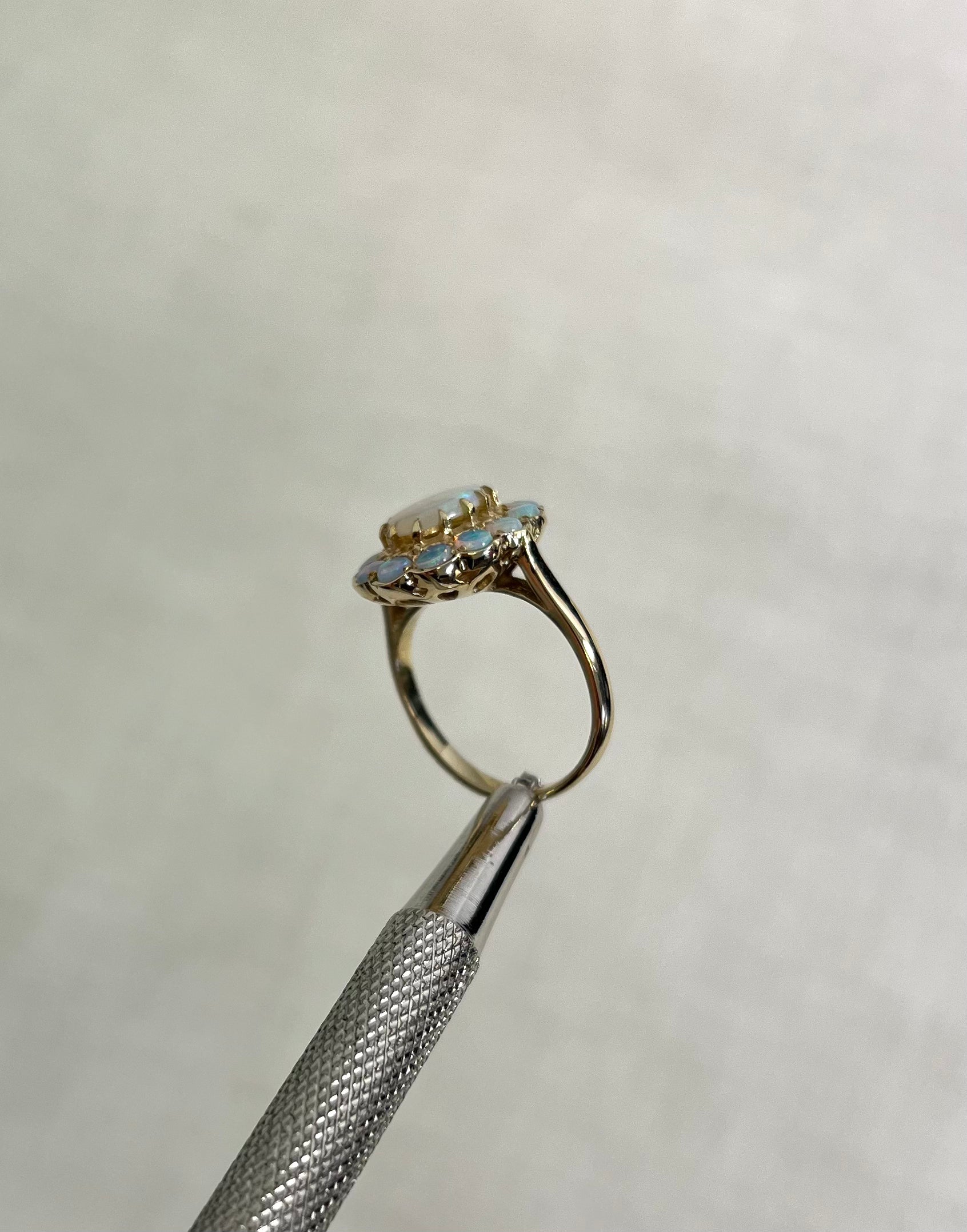 side view of gold ring with multiple opal stones, help by silver metal tool above white background