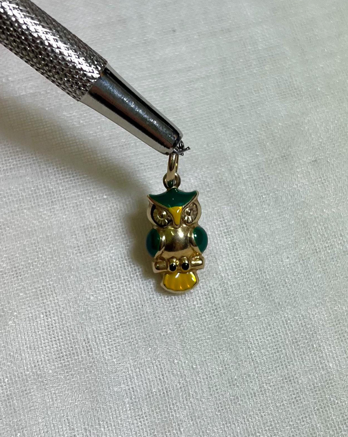 Gold owl charm with green and yellow accents held by tweezers on a light cream fabric background