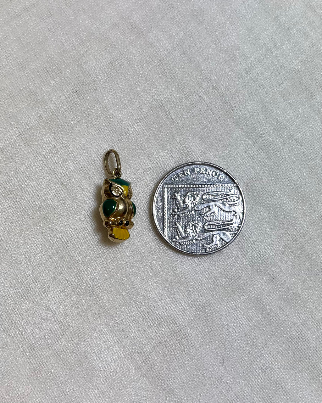 A small gold owl charm with green and yellow enamel details shown next to a silver ten pence coin for scale, on a light cream textured fabric background.