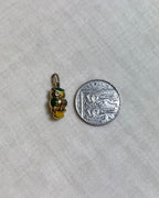 A small gold owl charm with green and yellow enamel details shown next to a silver ten pence coin for scale, on a light cream textured fabric background.
