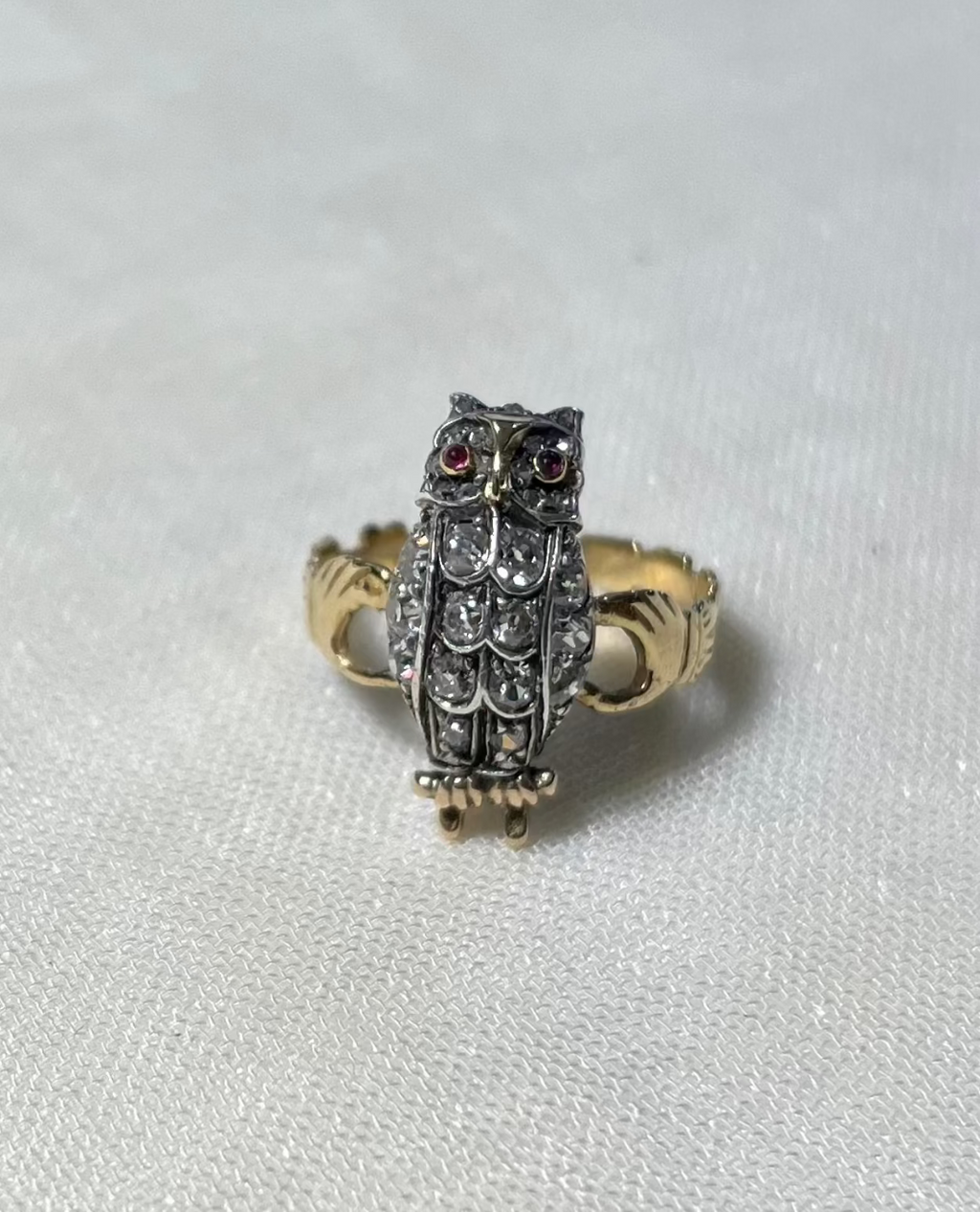 An owl-shaped ring featuring a silver-toned body with diamond stones, red gemstone eyes, and a gold-toned band, resting on a textured white fabric background.