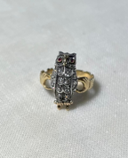 An owl-shaped ring featuring a silver-toned body with diamond stones, red gemstone eyes, and a gold-toned band, resting on a textured white fabric background.