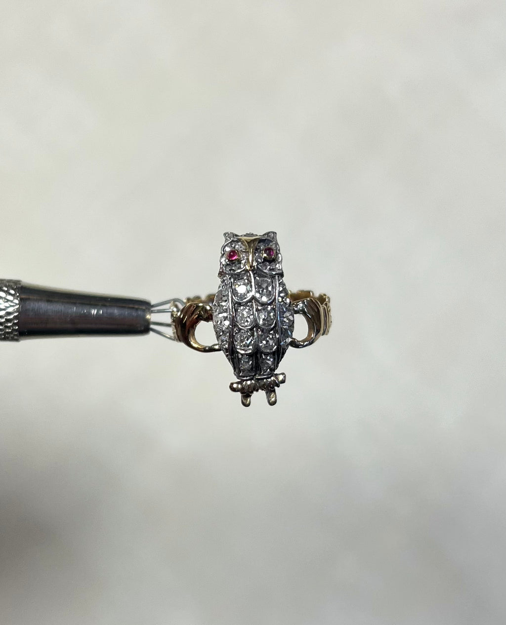 A small owl ring with red gemstone eyes and a textured feather pattern set with diamonds, held by a metal tweezer against a soft white background.