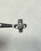 A small owl ring with red gemstone eyes and a textured feather pattern set with diamonds, held by a metal tweezer against a soft white background.
