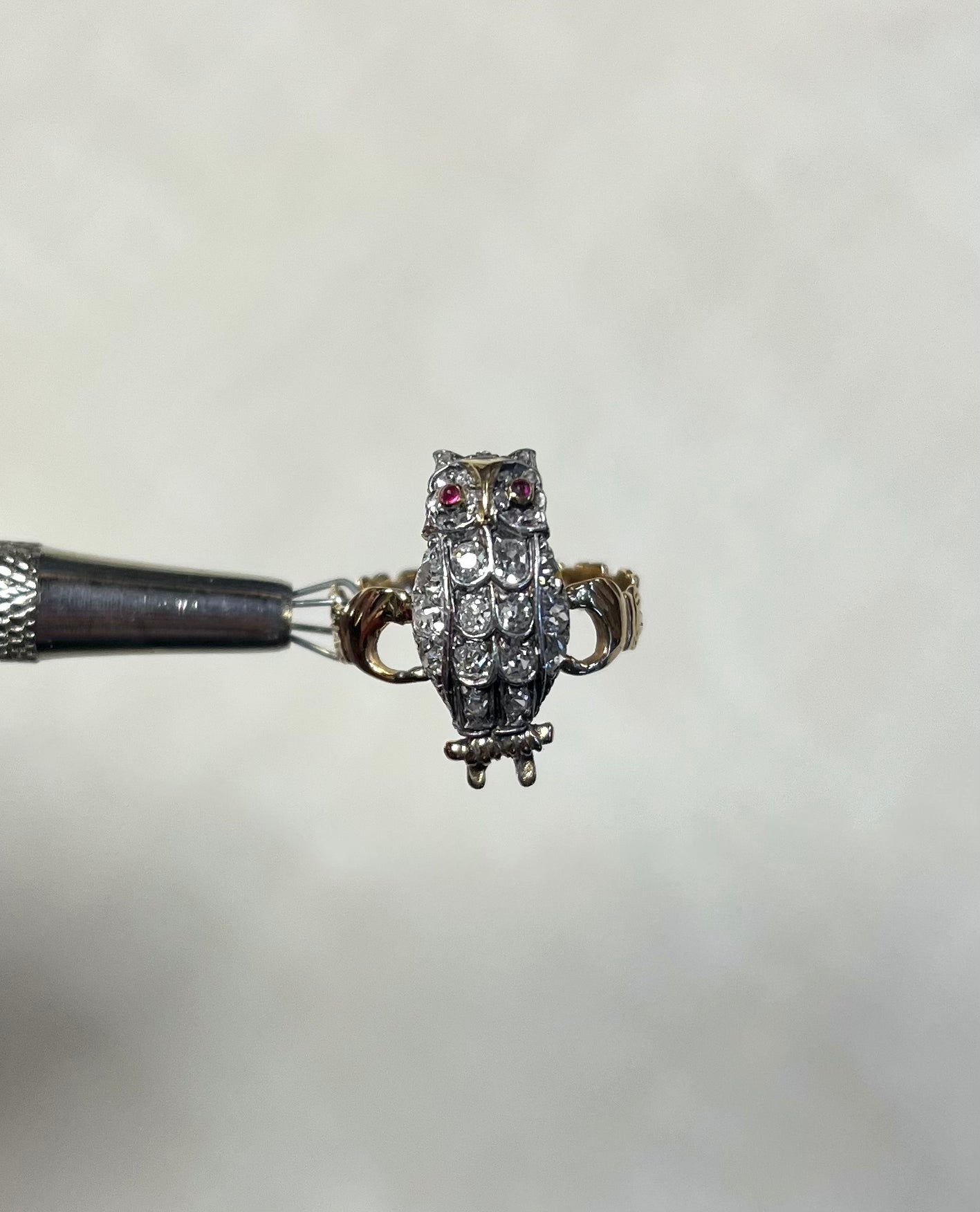 A small owl ring with red gemstone eyes and a textured feather pattern set with diamonds, held by a metal tweezer against a soft white background.