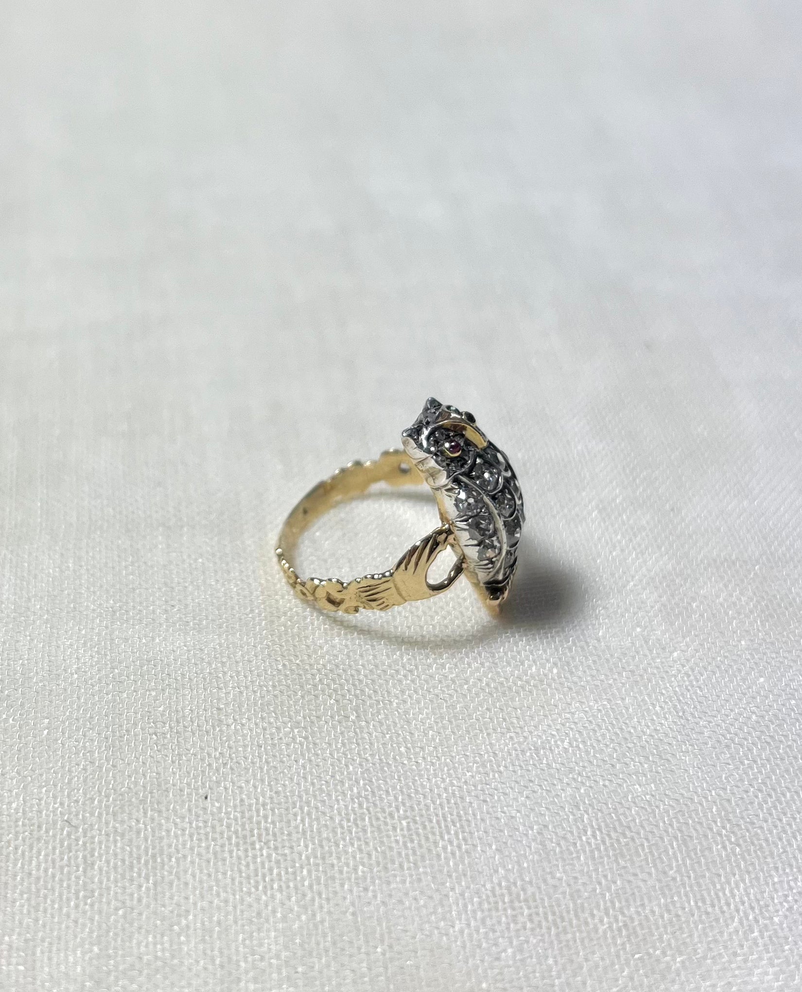 side view of gold owl ring set with diamonds and ruby eyes, on white fabric background