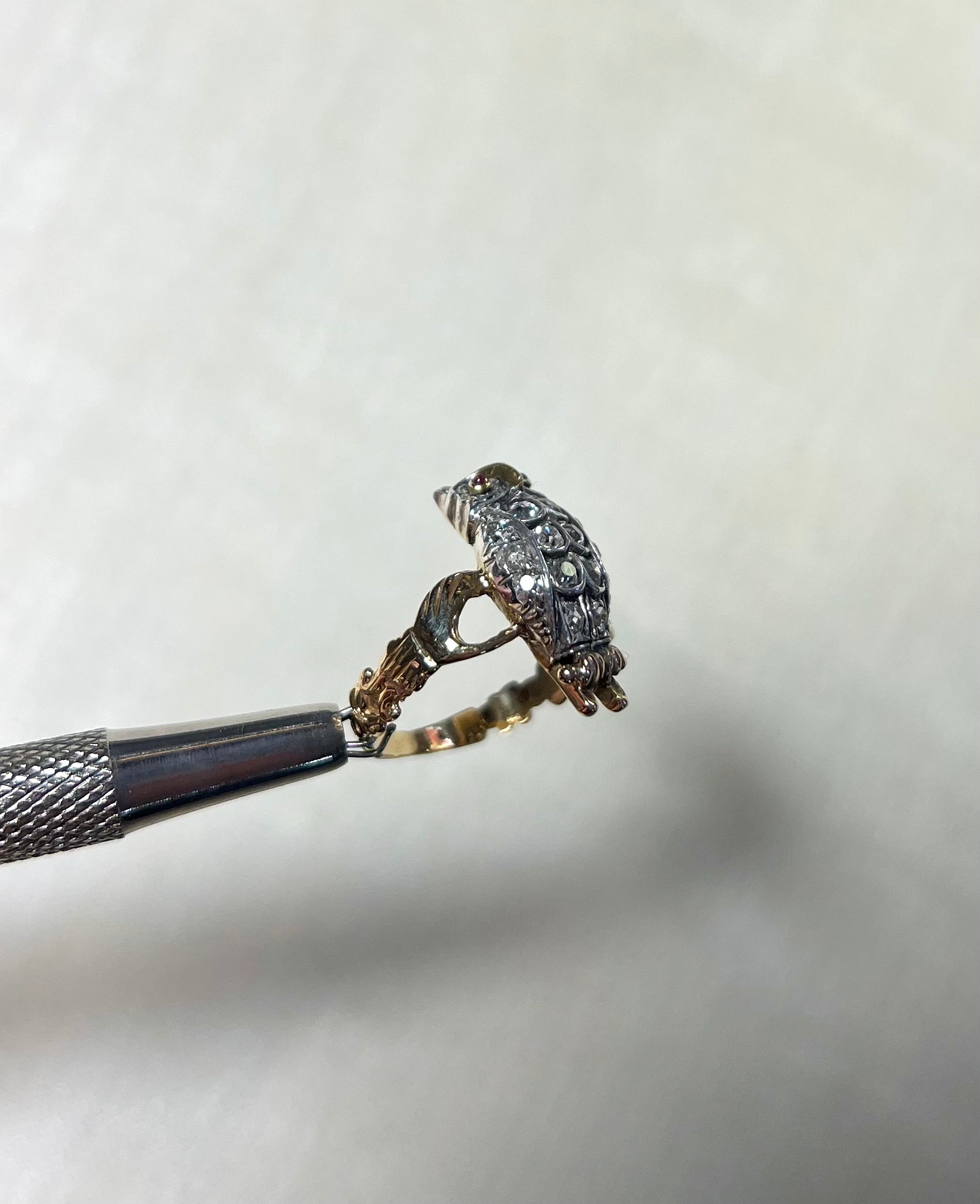 side view of bird shaped ring set with diamonds, held by silver metal tool against soft white background