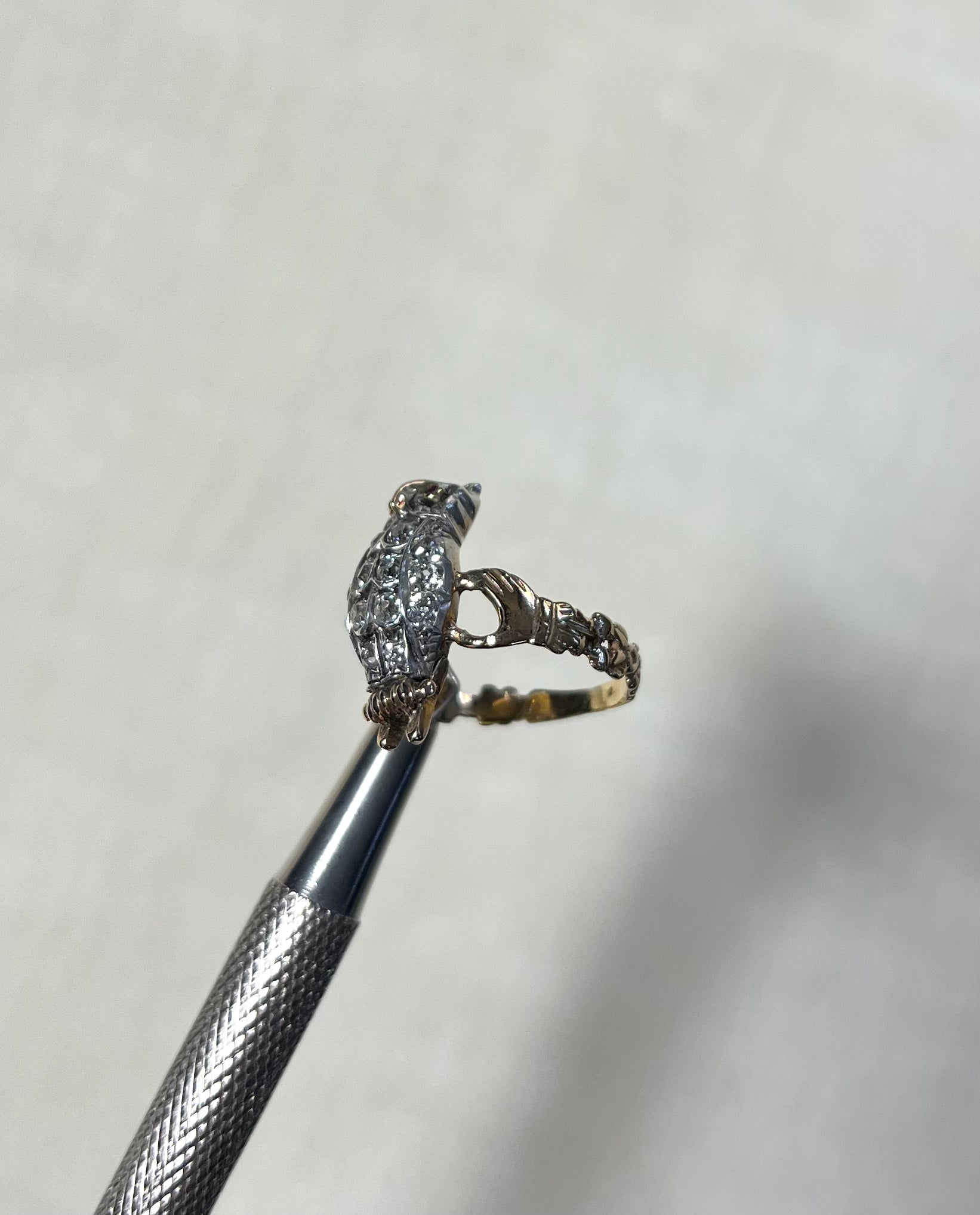 A small bird-shaped ring with a sparkling diamonds and a gold  band, held on a metal tweezer against a soft white background.