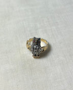 An owl-shaped ring with a textured silver body, small red gemstone eyes, and gold-toned side details, resting on a light beige fabric background.