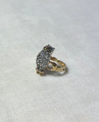 A vintage owl ring featuring a silver-toned owl body set with clear diamonds, small red gemstone eyes, and a gold band, resting on a textured off-white fabric background.