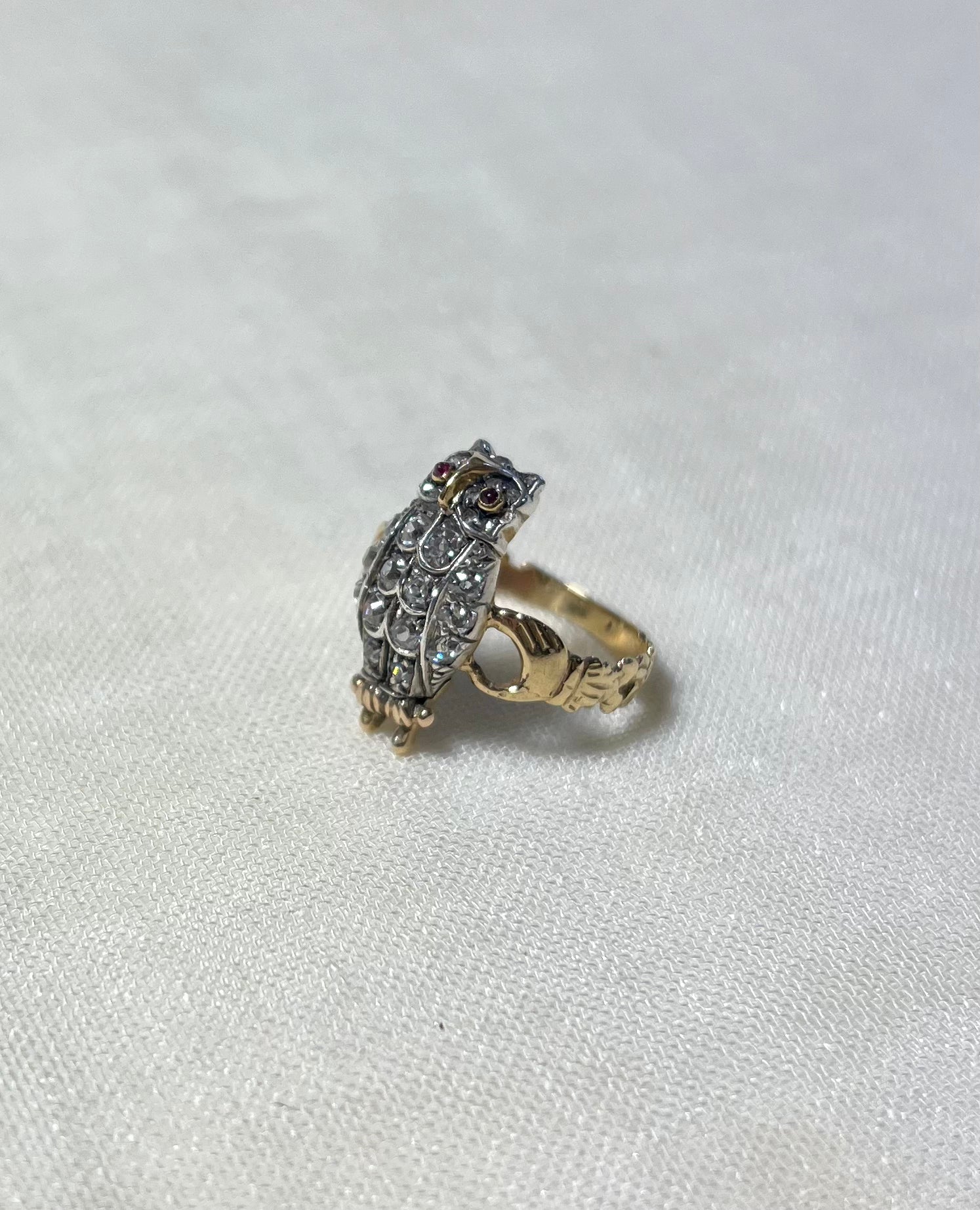 A vintage owl ring featuring a silver-toned owl body set with clear diamonds, small red gemstone eyes, and a gold band, resting on a textured off-white fabric background.