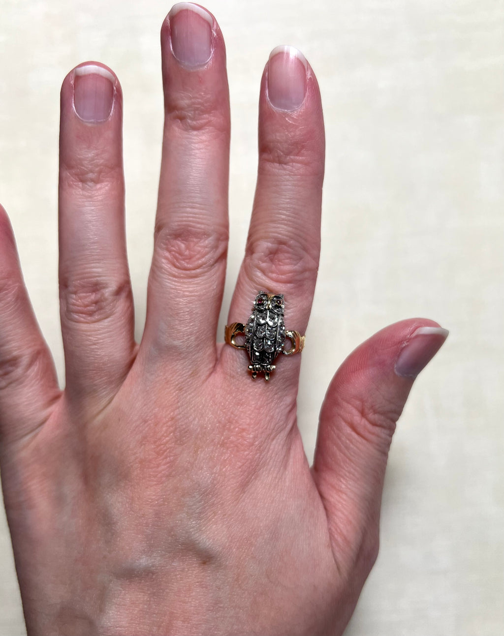 A close-up of a person's hand wearing a vintage owl-shaped ring. The ring features diamond set body and gold band, set against a plain light beige background.