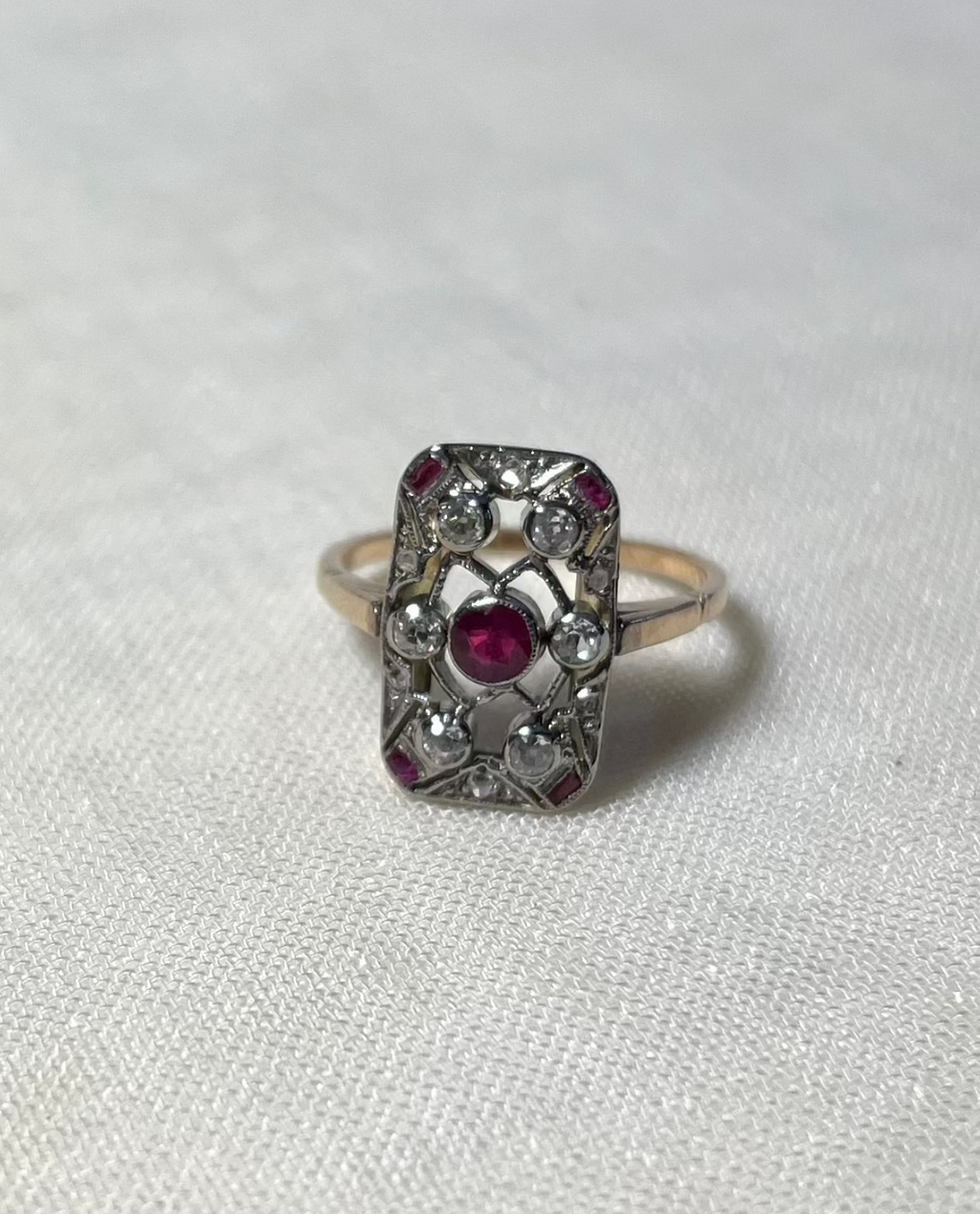 an antique gold ring with rectangular head set with centre ruby surrounded by an array of diamonds and rubies, resting on white fabric background