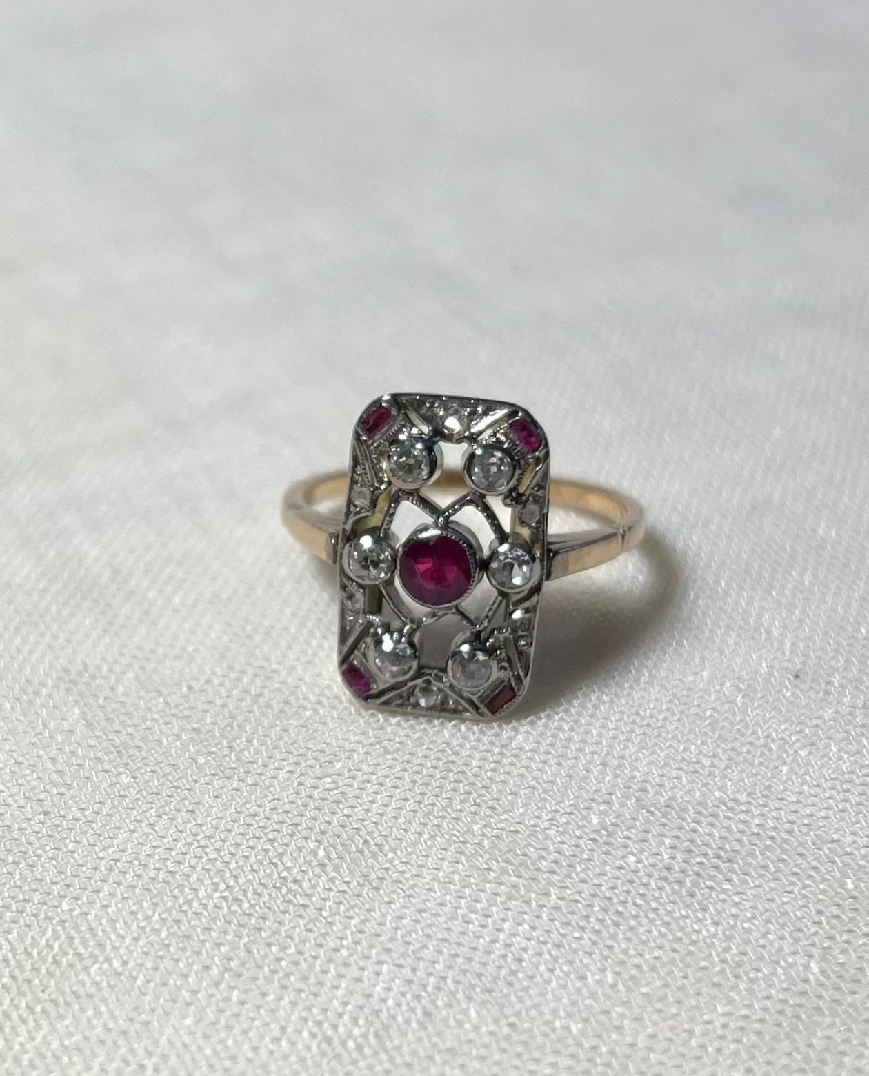 an antique gold ring with rectangular head set with centre ruby surrounded by an array of diamonds and rubies, resting on white fabric background
