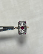 An ornate vintage-style ring with a rectangular shape and set with centre ruby and surrounding clear diamond  stones, held horizontally by a metal tool against a soft white fabric background.