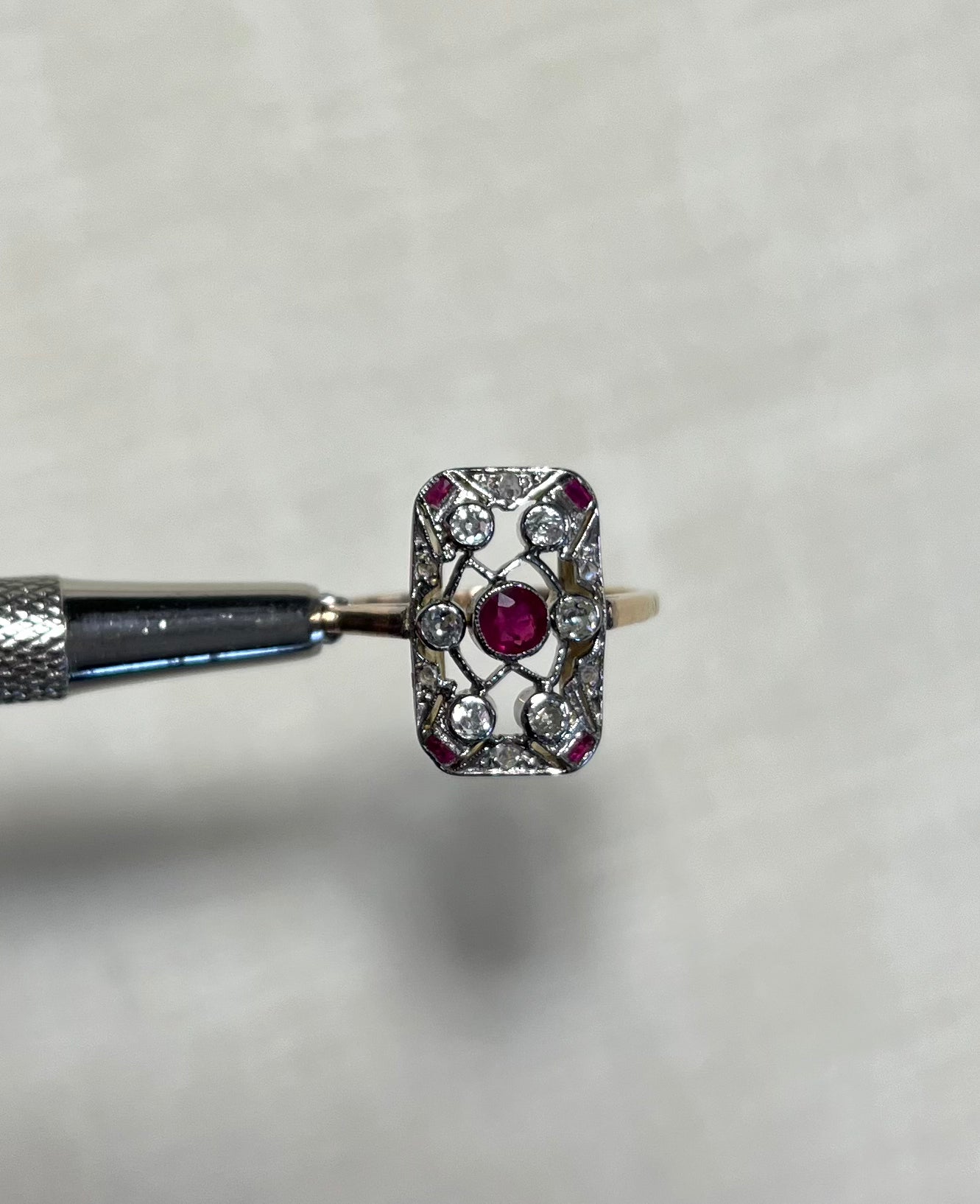 An ornate vintage-style ring with a rectangular shape and set with centre ruby and surrounding clear diamond  stones, held horizontally by a metal tool against a soft white fabric background.