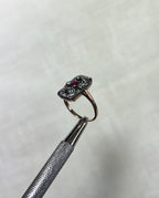 an antique gold ring with rectangular head set with rubies and diamonds, held by silver metal tool above soft white background 