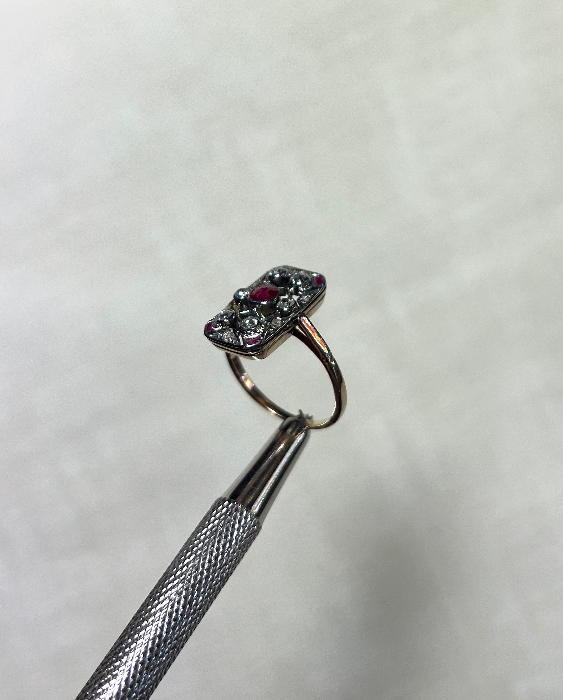 an antique gold ring with rectangular head set with rubies and diamonds, held by silver metal tool above soft white background 