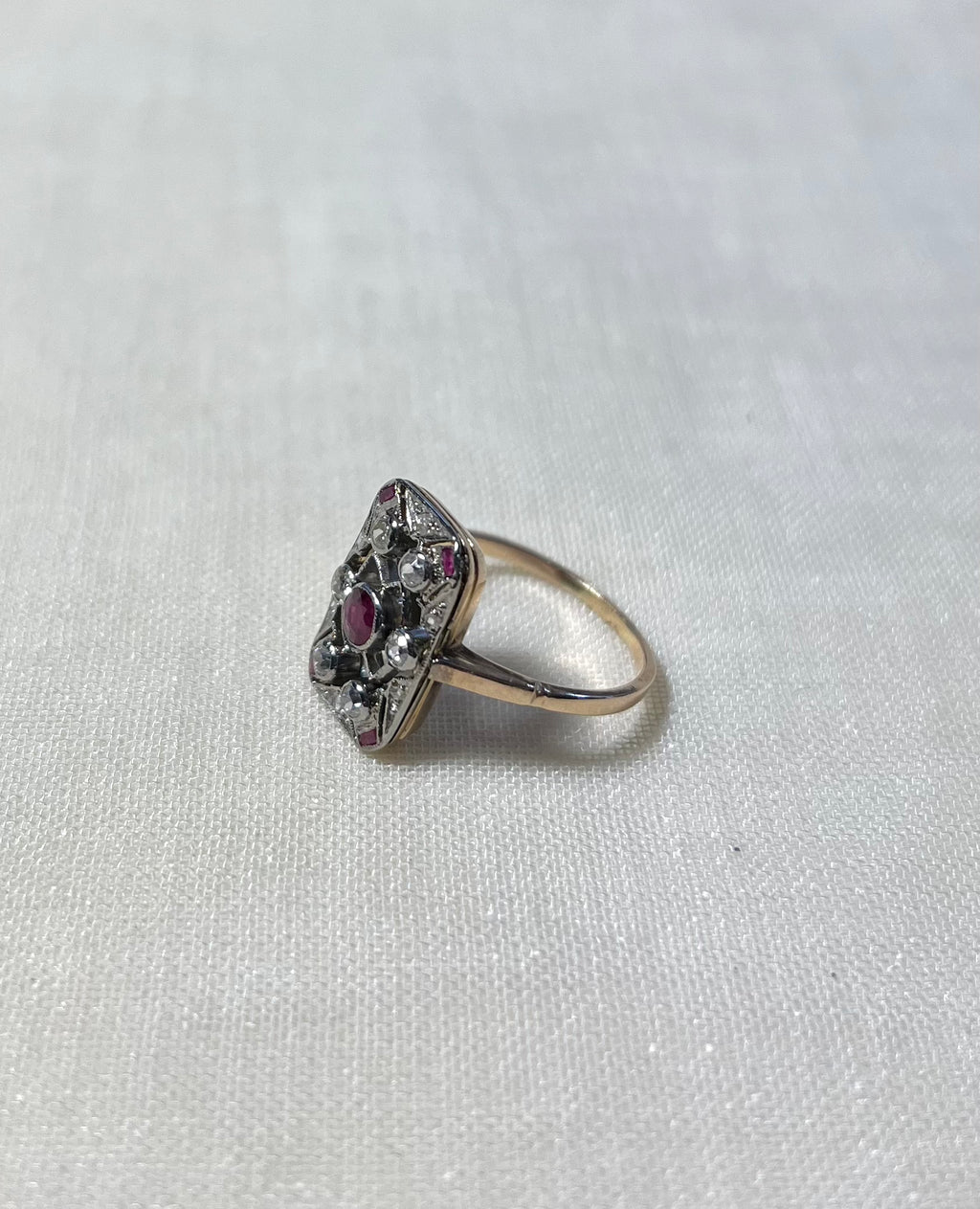 A vintage-style ring with a gold band and a decorative cluster setting featuring clear stones and a central pinkish-red gemstone, resting on a textured off-white fabric background.