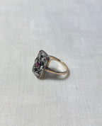 A vintage-style ring with a gold band and a decorative cluster setting featuring clear stones and a central pinkish-red gemstone, resting on a textured off-white fabric background.