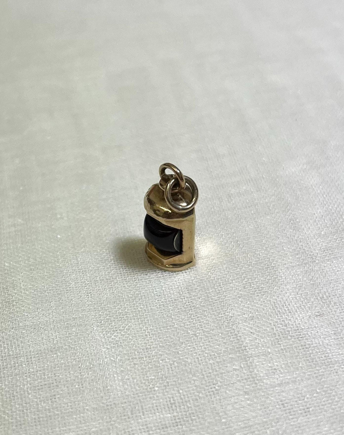 Small gold charm with a dark red center in the shape of ship lantern on a white fabric background