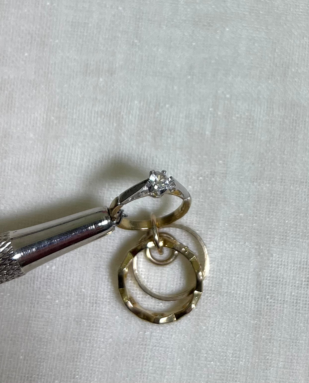 a close up of clear cubic zirconia set gold ring charm held by metal tool above white fabric background