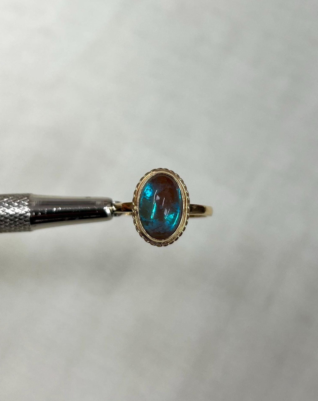 gold ring with iridescent blue stone, held by metal tool against light beige background