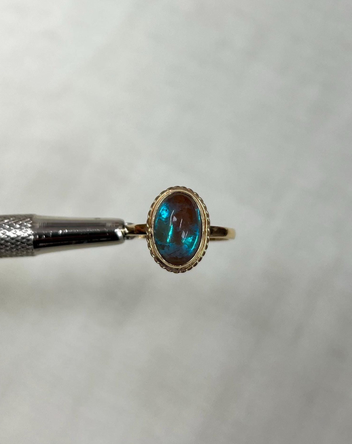 gold ring with iridescent blue stone, held by metal tool against light beige background