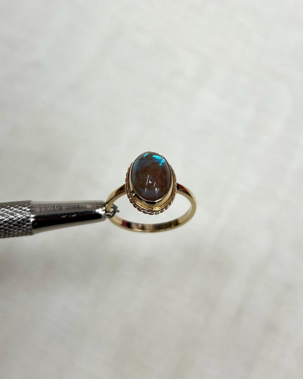 Gold ring with an oval gemstone held by tweezers on a light beige background