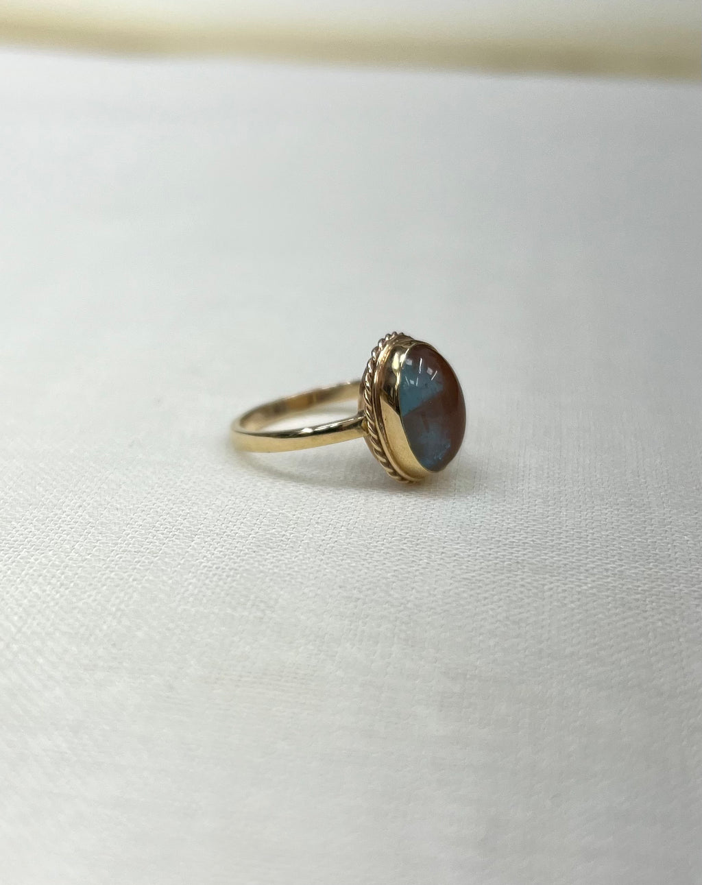 side view of gold ring with an oval cabochon stone showing blue and brown iridescent tones, resting on a textured white fabric surface.