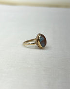 side view of gold ring with an oval cabochon stone showing blue and brown iridescent tones, resting on a textured white fabric surface.