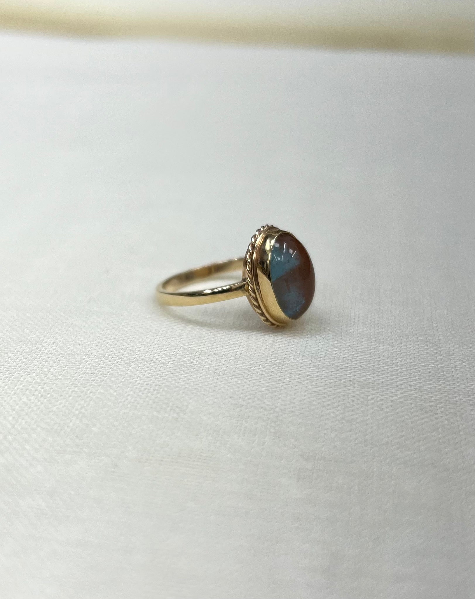 side view of gold ring with an oval cabochon stone showing blue and brown iridescent tones, resting on a textured white fabric surface.