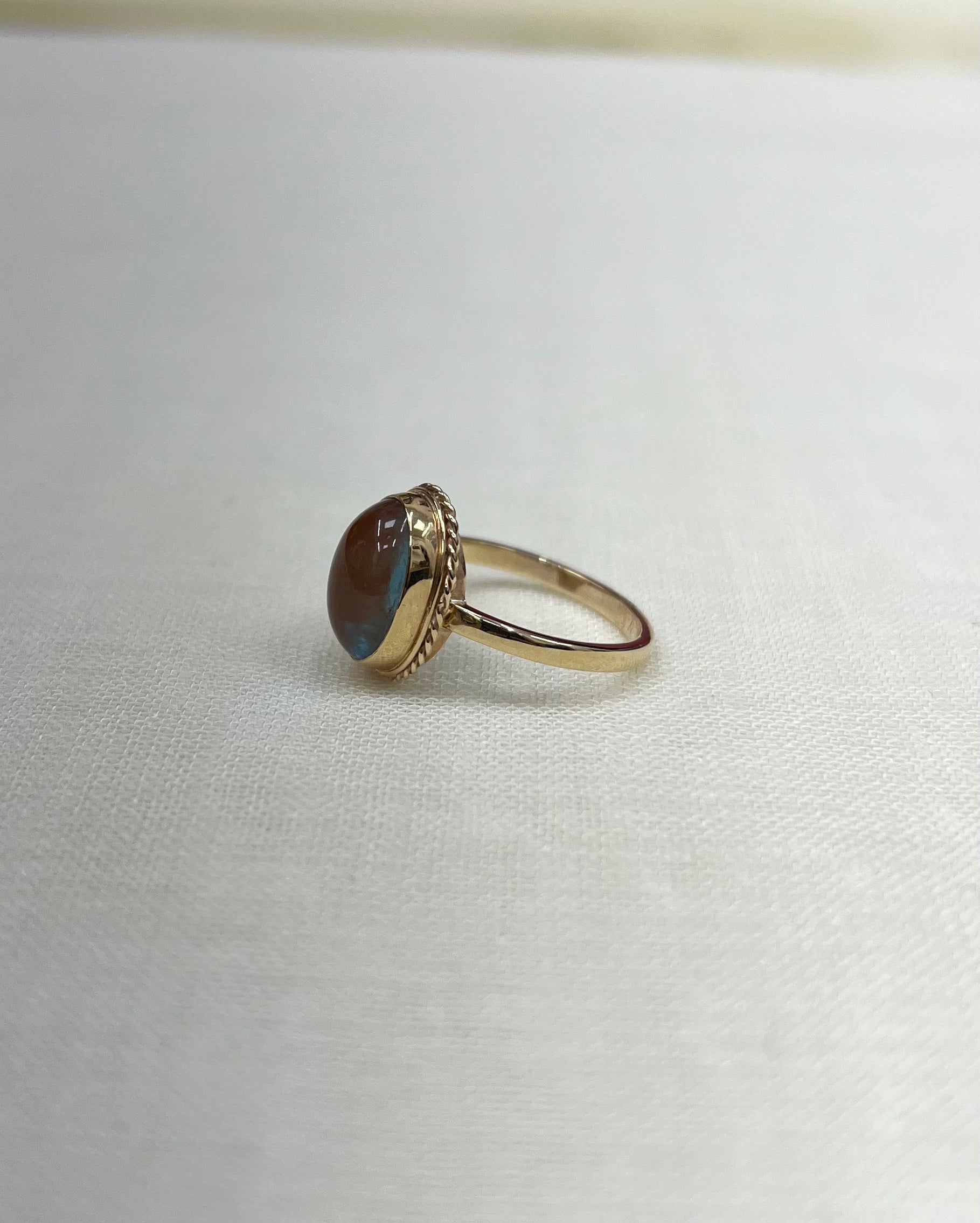 A gold ring with an oval brown and blue iridescent cabochon gemstone, resting on a textured white fabric surface.