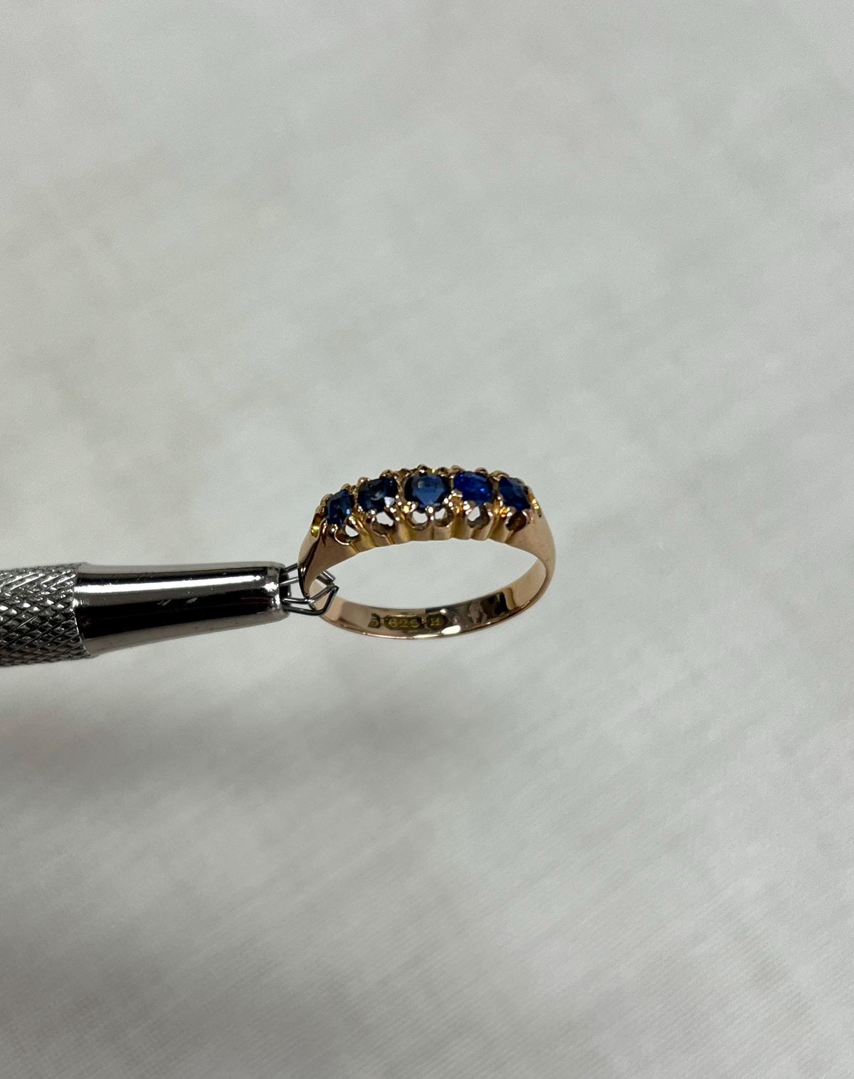 Gold ring with blue stones being held by silver tool on beige background