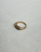 A gold ring with a row of small dark blue gemstones, photographed on a light cream coloured textured fabric background.
